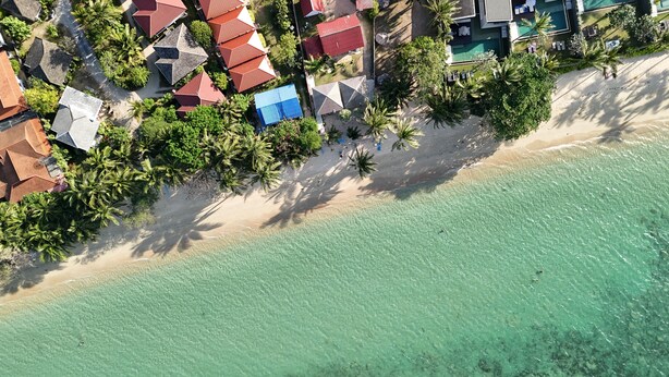 An aerial view of a coastal area with a beach, clear turquoise water, and a row of buildings with red roofs. - Ocean Peek Villa-Bang Po  (Chang Wat Surat Thani)