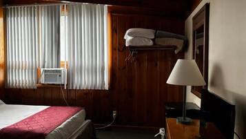 Habitación de hotel con cama, escritorio con lámpara, calentador de pared y ventana con persianas.