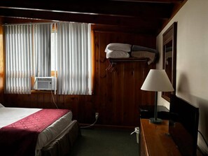 A hotel room with a bed, a desk with a lamp, a wall-mounted heater, and a window with blinds. - Cozy 1-bedroom hotel room, Hamilton - #350 Oneida (Hamilton)