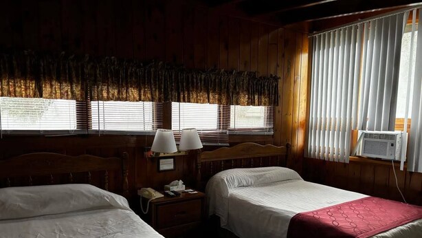 Una habitación con dos camas, una mesita de noche, una lámpara y una ventana con persianas.
