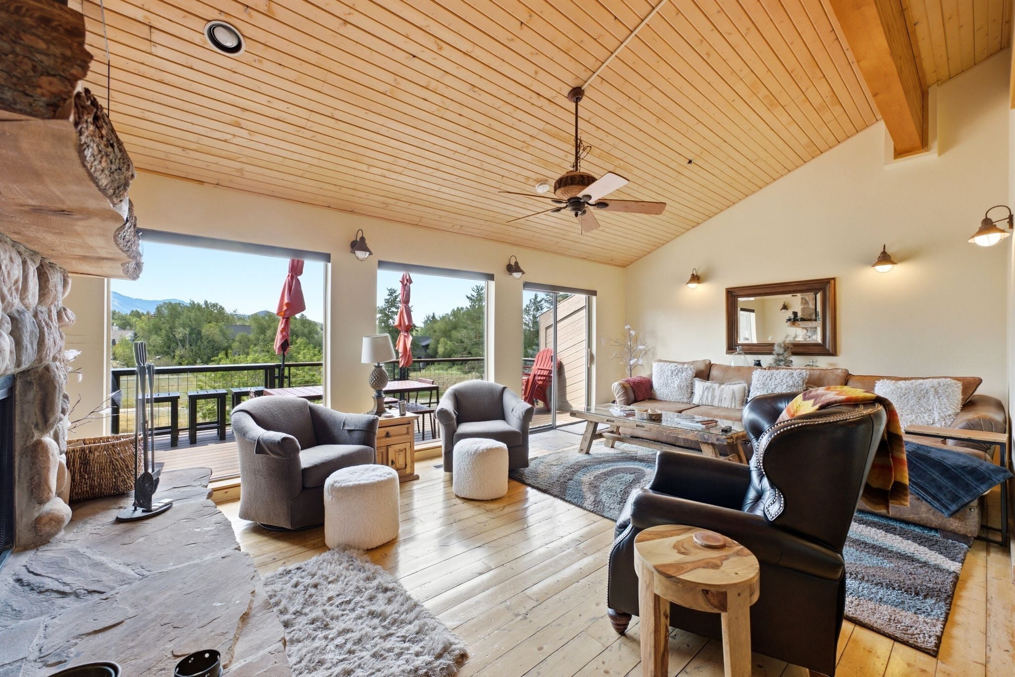 A spacious living area with a high ceiling, wooden flooring, and a fireplace. There are multiple seating options including a sofa, armchairs, and ottomans. The room features large windows offering a view of the outdoors.