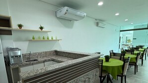 A modern kitchen with granite countertops, a built-in sink, and a dining area with round tables and chairs. - Luxor Tambau (João Pessoa)