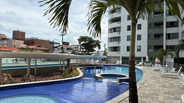 A swimming pool area with a palm tree, lounge chairs, and a building in the background. - Luxor Tambau (João Pessoa)