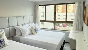 A hotel room with two beds, a desk, and a window with a view of buildings and parked cars. - Luxor Tambau (João Pessoa)