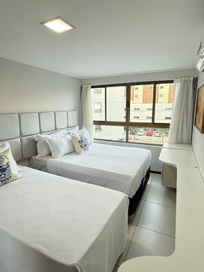 A hotel room with two beds, a desk, and a window with a view of buildings and parked cars.