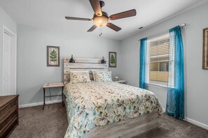 A bedroom with a bed, a nightstand, a wooden dresser, a ceiling fan, and a window with curtains. - Drift Away Pet Friendly Beach Side Town Home (Surfside)