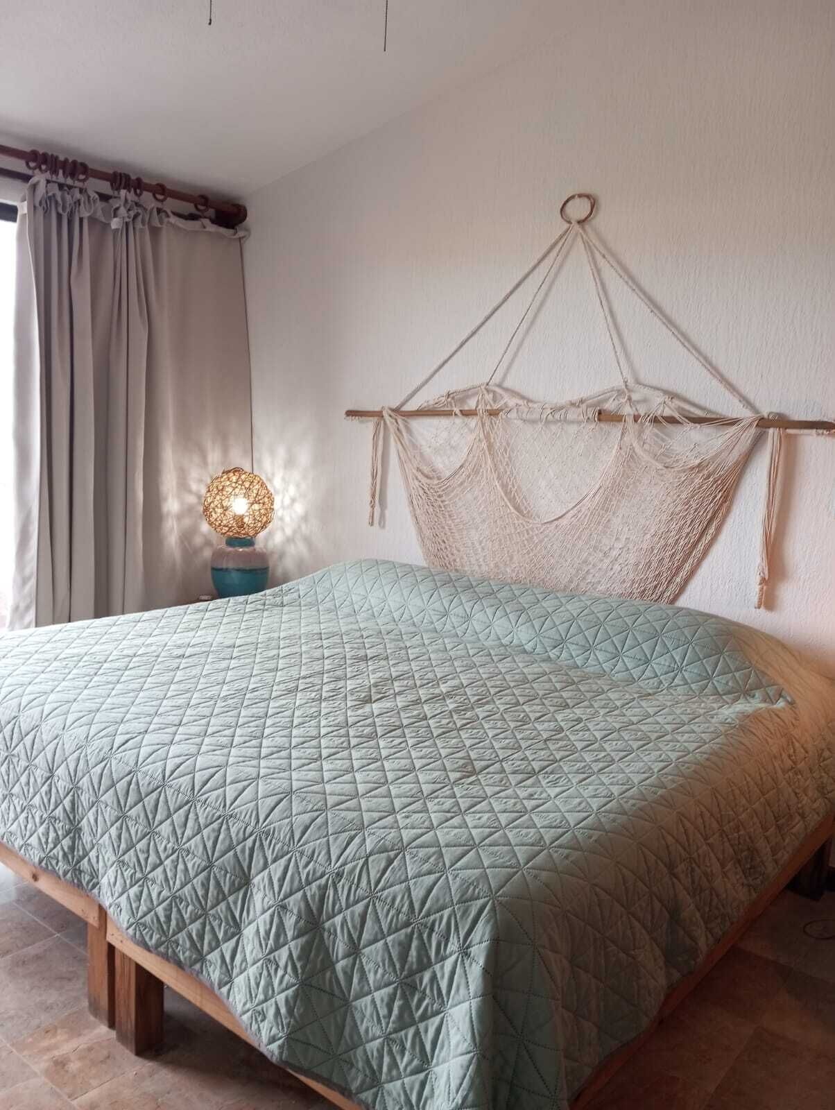 Un letto con una trapunta a motivi geometrici, una testiera in legno con decorazioni in macramè, una lampada da comodino e una finestra con tende.
