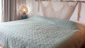 Ein Bett mit geometrischem Patchworkmuster, ein Holzkopfbrett mit Makramee, eine Nachttischlampe und ein Fenster mit Vorhängen.