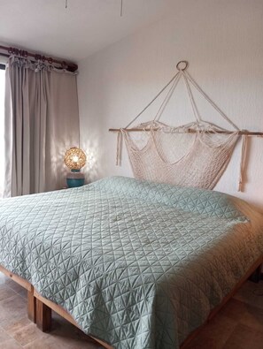 Ein Bett mit geometrischem Patchworkmuster, ein Holzkopfbrett mit Makramee, eine Nachttischlampe und ein Fenster mit Vorhängen.
