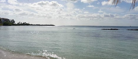 Una playa con agua cristalina, una orilla de arena y una costa lejana con edificios y vegetación.