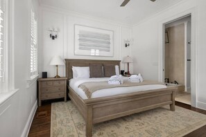 Ein Schlafzimmer mit einem hölzernen Bett, zwei Nachttischen, einem Schreibtisch und einem großen Fenster mit Jalousien, sowie einer Tür zu einem anderen Raum.
