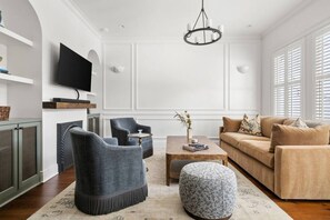 A living room with a television, two armchairs, a sofa, a coffee table, and a patterned ottoman. - Luxury 6BR w/ Pool Downtown CHS (Charleston)