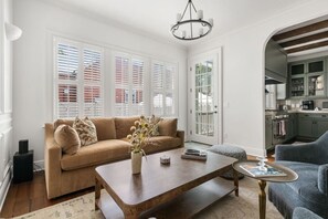 A living room with a sofa, coffee table, and a blue armchair. There is a kitchen visible through an archway. - Luxury 6BR w/ Pool Downtown CHS (Charleston)