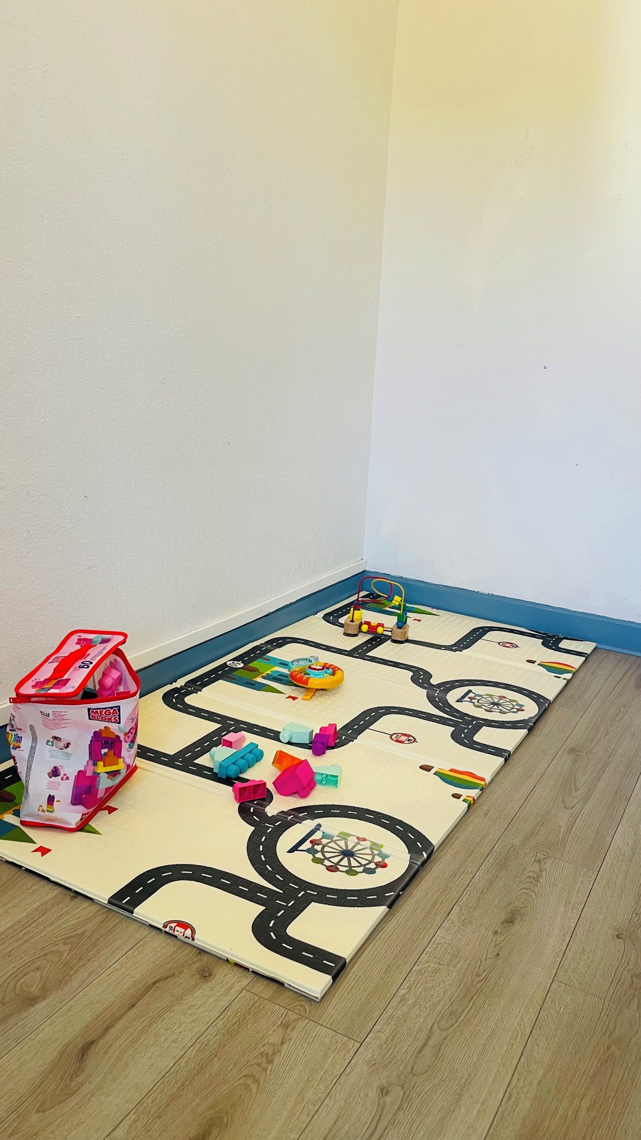 A room with a wooden floor and a colorful play mat featuring a road and various toys.