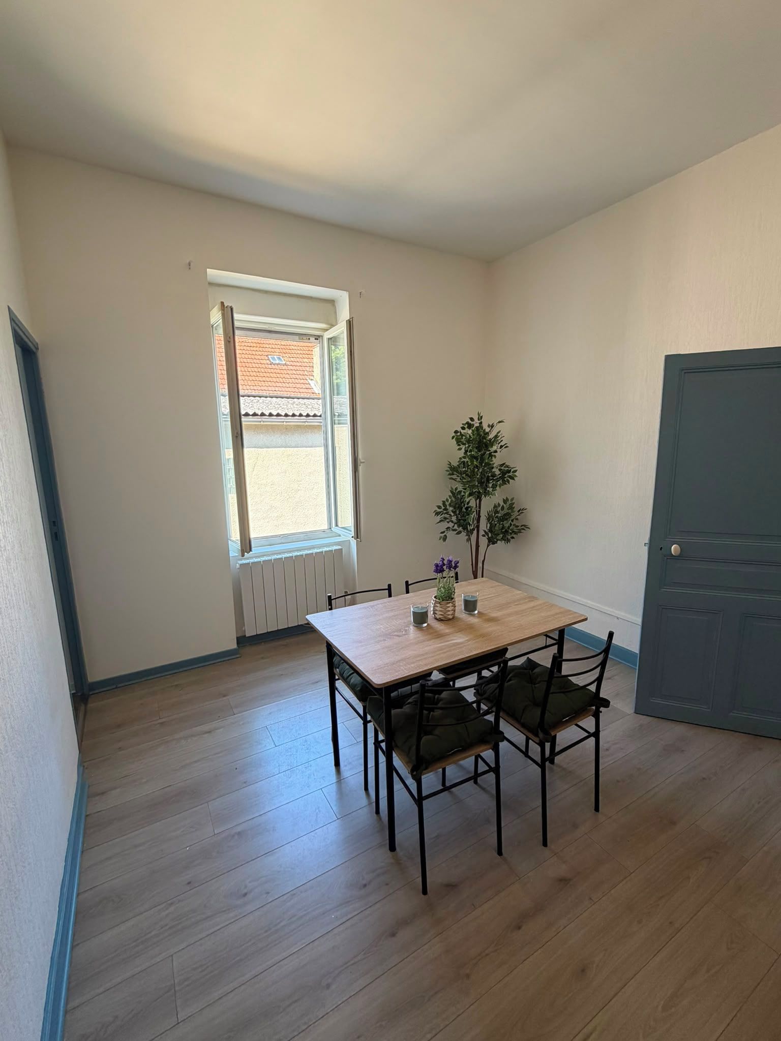 Een kamer met een houten tafel, vier stoelen, een raam met uitzicht op een gebouw, een radiator en een deur.