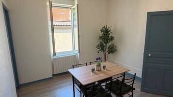 Een kamer met een houten tafel, vier stoelen, een raam met uitzicht op een gebouw, een radiator en een deur.