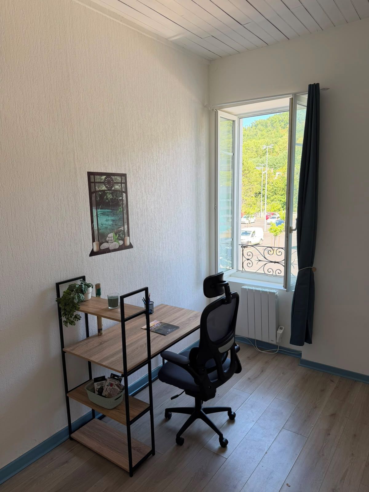 Un pequeño espacio de oficina con un escritorio, una silla y estanterías. Hay una ventana con cortinas y vistas al exterior.