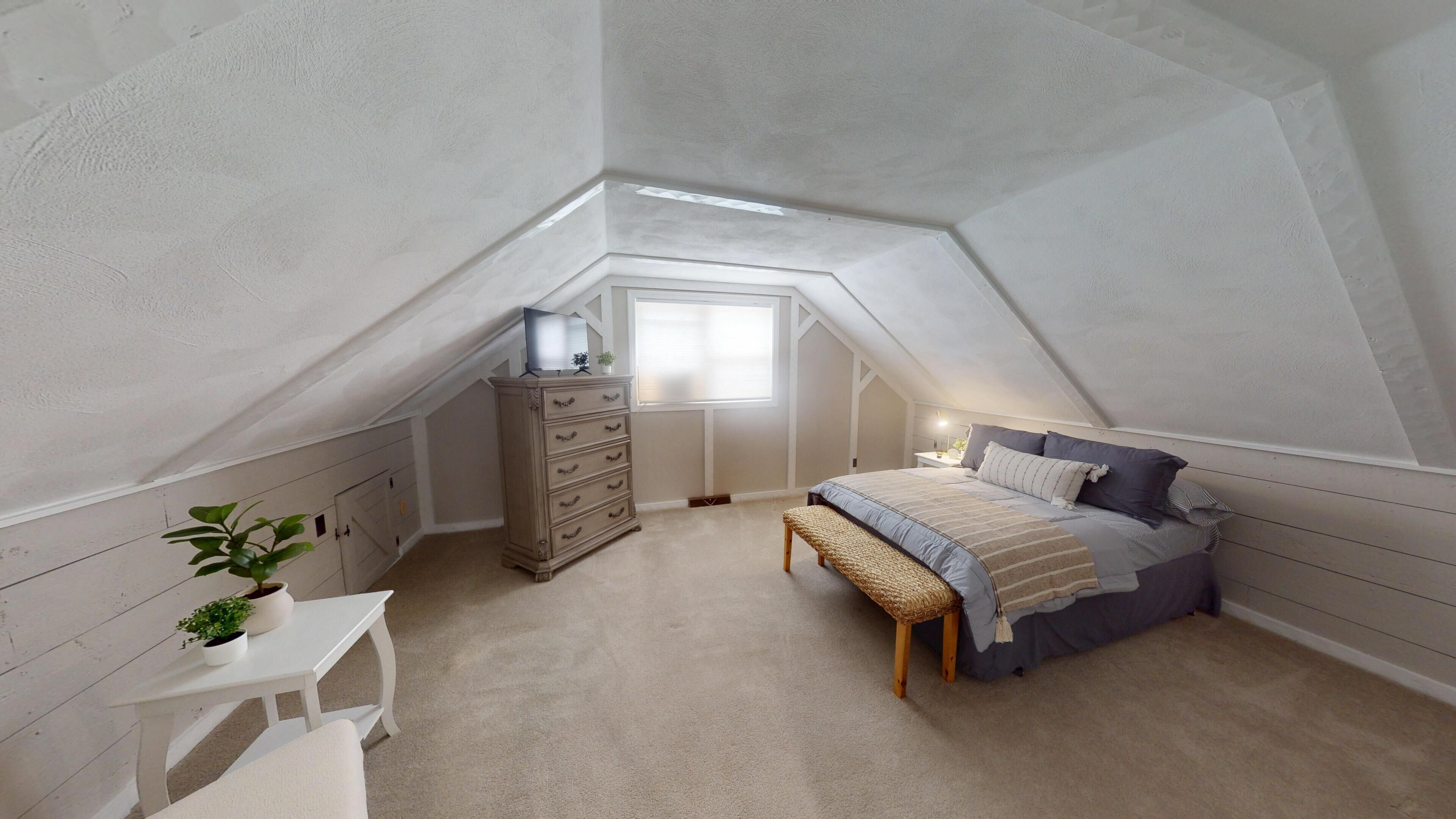 Una camera da letto con soffitto spiovente, un letto, una cassettiera, un tavolino e una finestra.