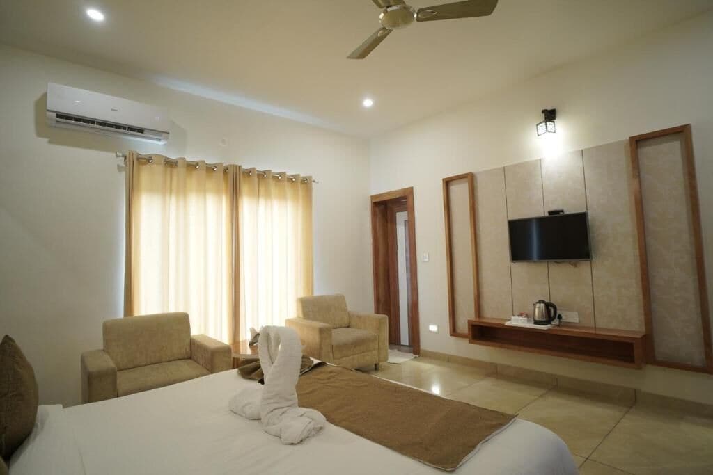 A hotel room with a bed, two armchairs, a television on the wall, and an air conditioning unit.