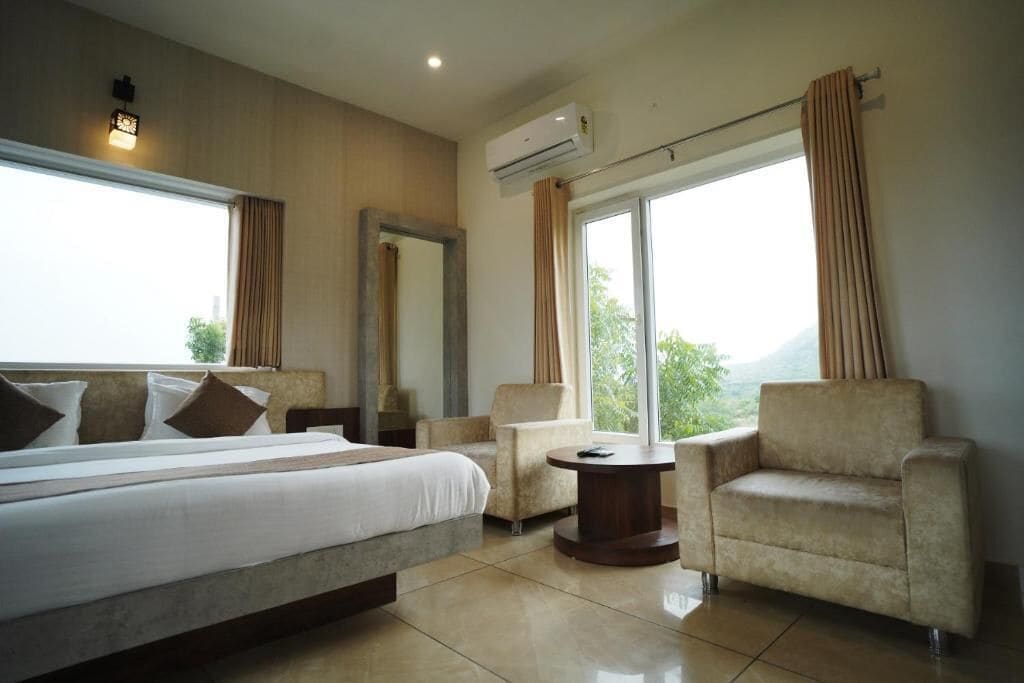 Habitación de hotel con cama, dos sillones, una mesita y un gran ventanal con cortinas.