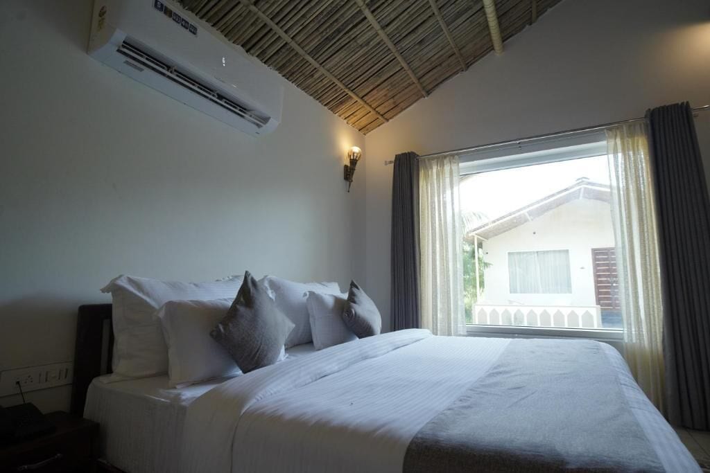 A hotel room with a bed, an air conditioning unit, a window with curtains, and a view of a building outside.