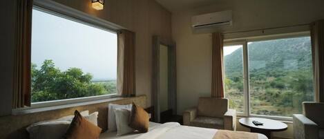A hotel room with a large bed, a sitting area with a table, and a view of greenery through large windows.