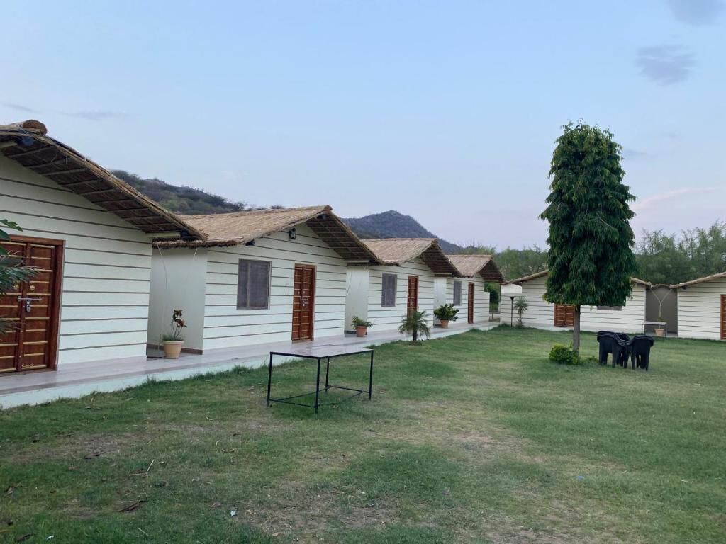 A row of white cabins with brown doors and windows, a grassy area, and a large tree.