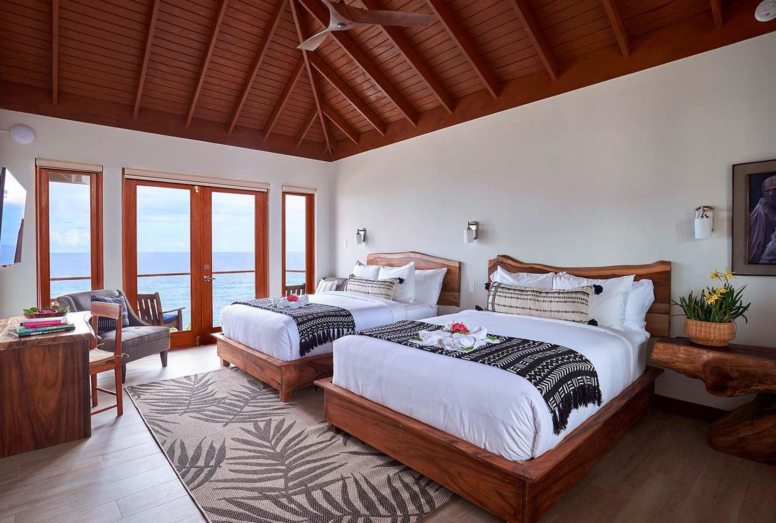 A hotel room with two beds, a desk, and a chair. There are large windows with a view of the sea.