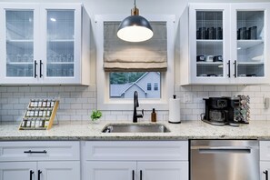A modern kitchen with white cabinetry, a stainless steel sink, a coffee maker, and a window with a view of a house.