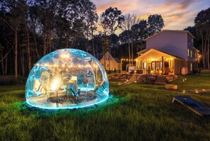 A transparent dome structure with interior lighting, a house with a porch, and a grassy area.
