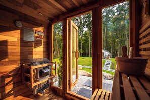 A sauna room with wooden walls, a sauna heater, and a view of a forested area through glass doors.