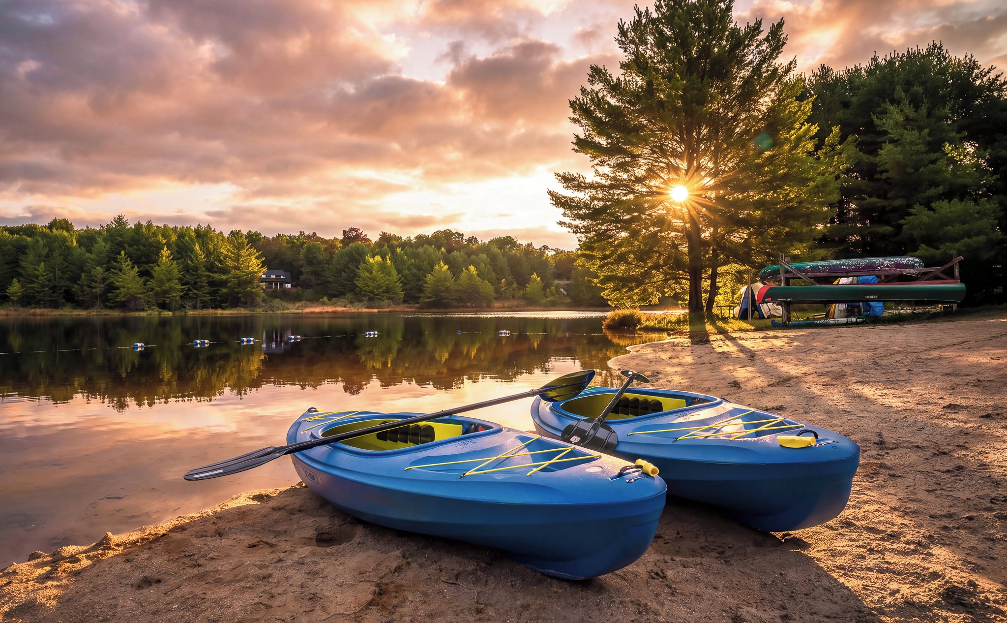 Two kayaks on a sandy shore by a calm lake with a forest in the background and a sunset.