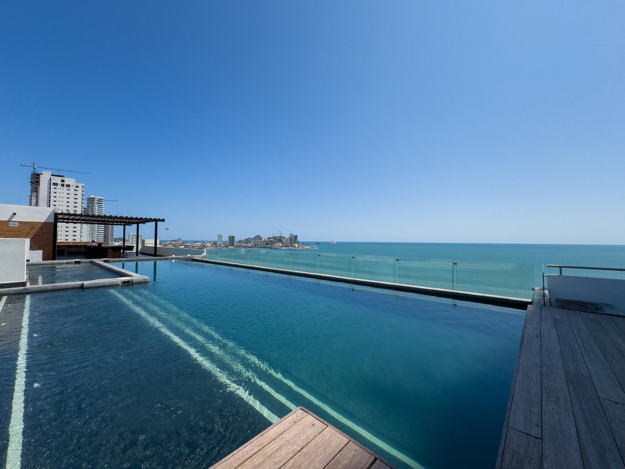 Une piscine à débordement avec une terrasse en bois, une eau d’un bleu cristallin et une vue sur l’océan et le paysage urbain lointain.