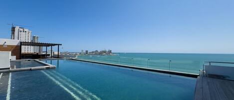 An infinity pool with a wooden deck, clear blue water, and a view of the ocean and distant cityscape.
