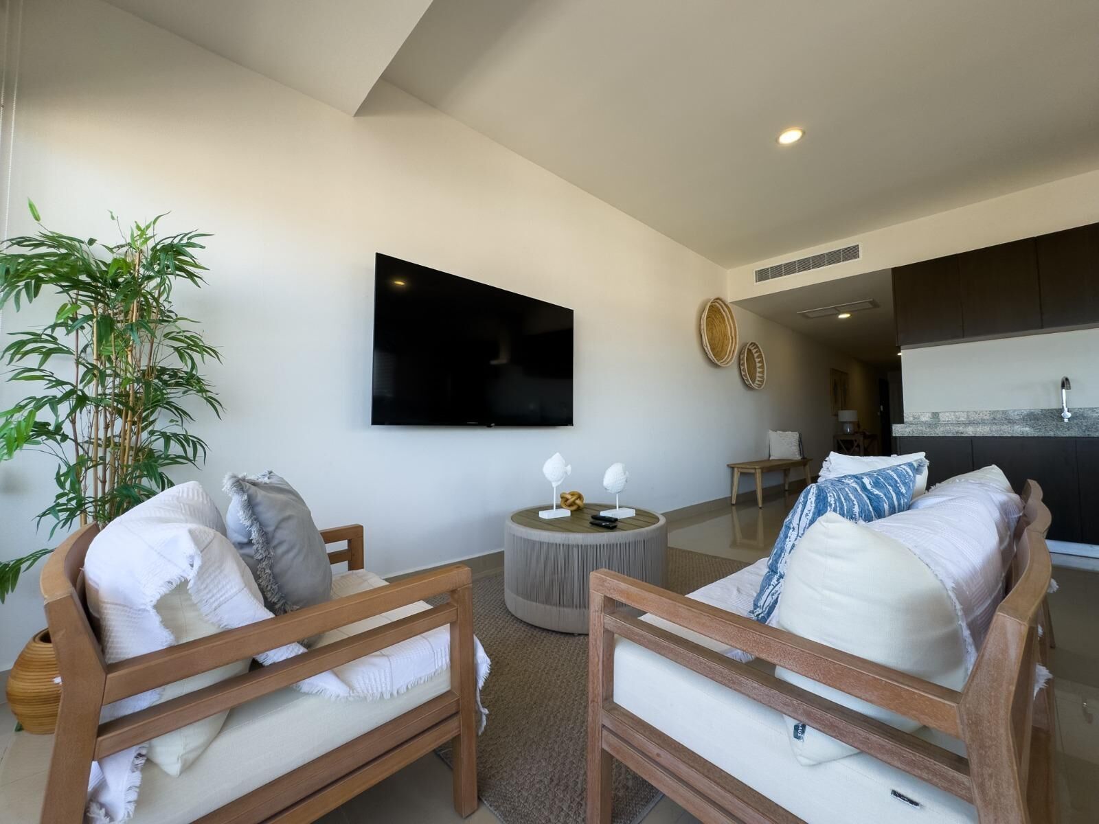 Uma sala de estar moderna com uma TV de tela plana na parede, duas cadeiras de madeira com almofadas, uma mesa redonda pequena com objetos decorativos e uma área de cozinha ao fundo.