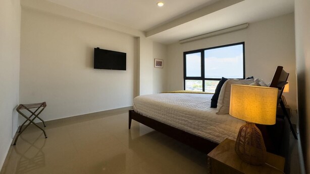 A bedroom with a bed, a television mounted on the wall, a window with a view, and a bedside lamp. - Spectacular Oceanview Escape in Torre 5 | Minutes to Centro! (Mazatlan)