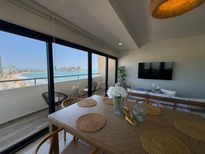 A dining area with a view of the beach and ocean, a flat-screen TV on the wall, and a wooden table with woven placemats. - Spectacular Oceanview Escape in Torre 5 | Minutes to Centro! (Mazatlan)