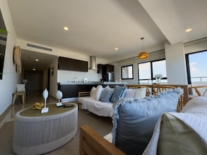 A modern living area with a sofa, a round coffee table, and a dining table with chairs. There is a kitchen area with cabinets and a window. - Spectacular Oceanview Escape in Torre 5 | Minutes to Centro! (Mazatlan)