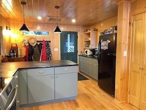 A kitchen with wooden cabinets, a black refrigerator, and a microwave. There are hanging hats and a door with a window.