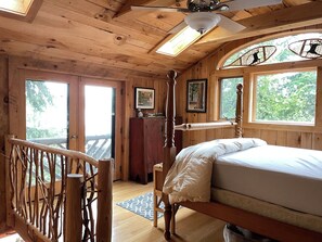 A wooden cabin bedroom with a four-poster bed, a ceiling fan, and a large window with a view of trees. - Lakefront 4-bedroom property with spectacular views of Blue Mountain Lake. (Blue Mountain Lake)