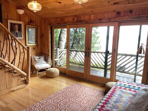 Interior de una cabaña de madera con cama, una silla de mimbre, una escalera de madera y un amplio ventanal corredero que da a una terraza exterior.
