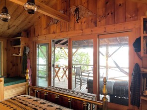 A wooden cabin interior with a bed, a window overlooking a deck, and a hanging light fixture.