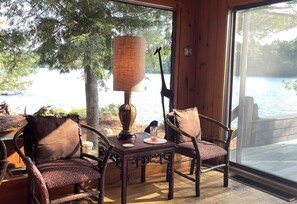 A wooden interior with a table, chairs, a lamp, and a view of a lake through a large window.