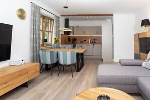 Ein modernes Wohnzimmer mit Sofa, Essbereich mit Stühlen, Küche mit Schränken und einem Fernseher links.