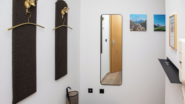 A hallway with a mirror, two wall-mounted hooks, a door, and a small shelf with a framed certificate.