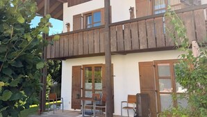A two-story building with a balcony, a table and chairs outside, and a grassy area in front.