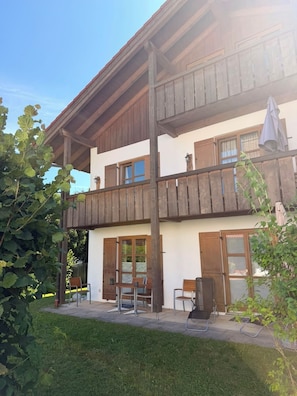 A two-story building with a balcony, a table and chairs outside, and a grassy area in front.
