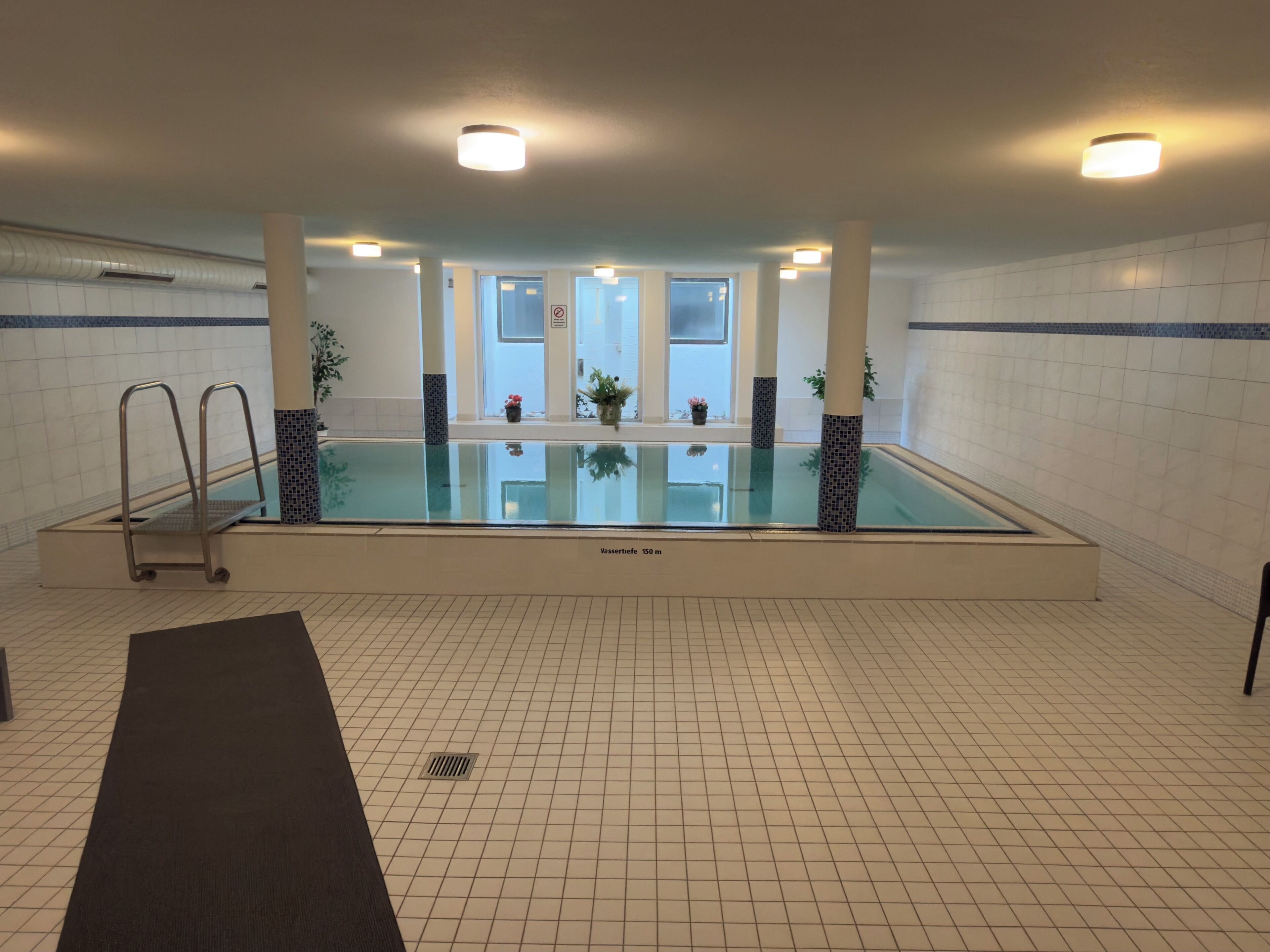 An indoor swimming pool with a diving board, tiled walls, and ceiling lights.