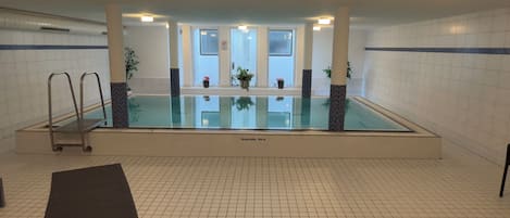 An indoor swimming pool with a diving board, tiled walls, and ceiling lights.