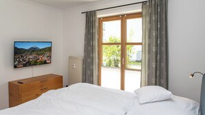 A bedroom with a bed, a wooden dresser, a radiator, a television on the wall, and a window with curtains.